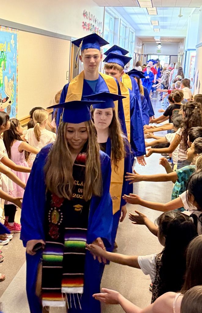 Graduates in caps and gowns walk down a hallway. Younger children reach out to touch the graduates' hands.