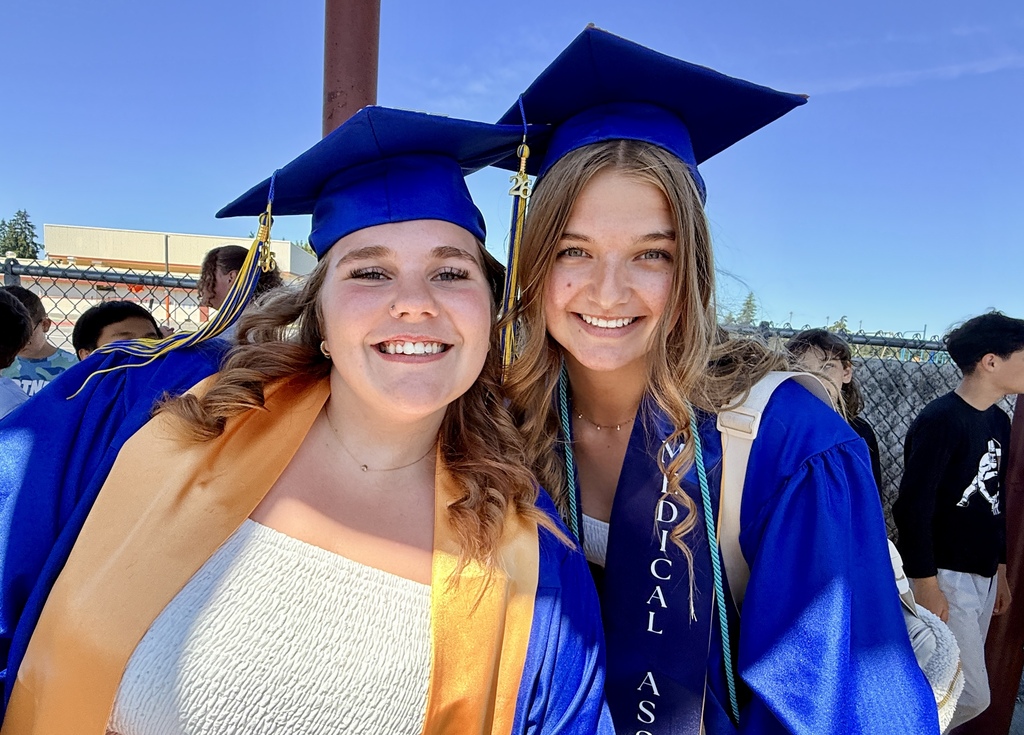 Two women, both in blue gowns and caps, stand smiling. One wears a sash, and the other a white shirt.