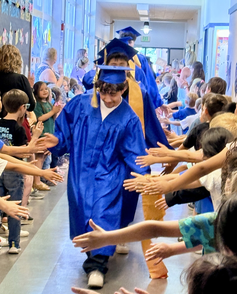 A young person in a blue graduation gown walks through a hallway, with children reaching out to greet them.