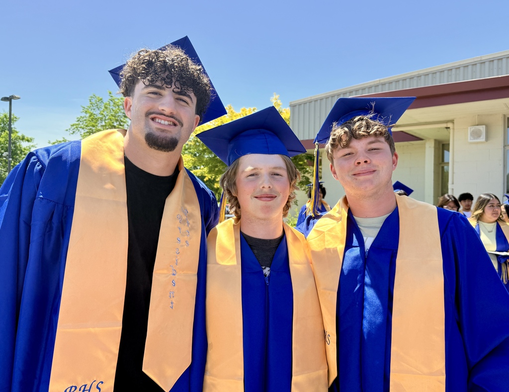 Three individuals wearing graduation gowns and caps stand in front of a building with a crowd of people.