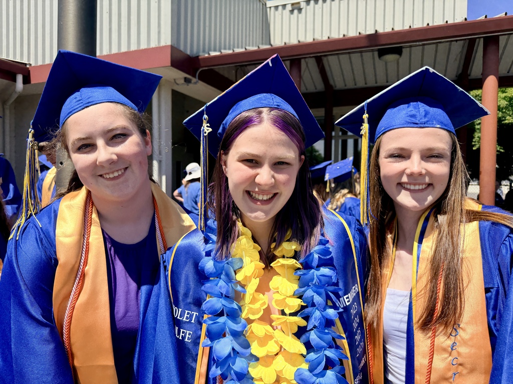 Three women in blue caps and gowns smile in front of a building. One wears a blue and yellow lei.