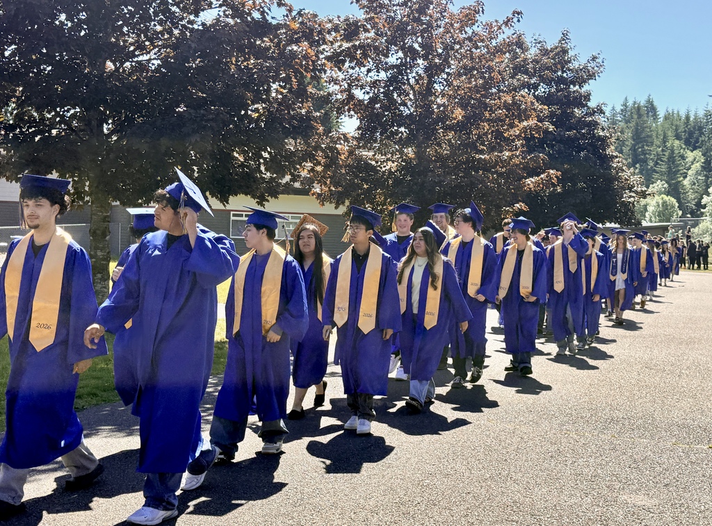 A line of people in blue caps and gowns walk on a path with trees and a building.