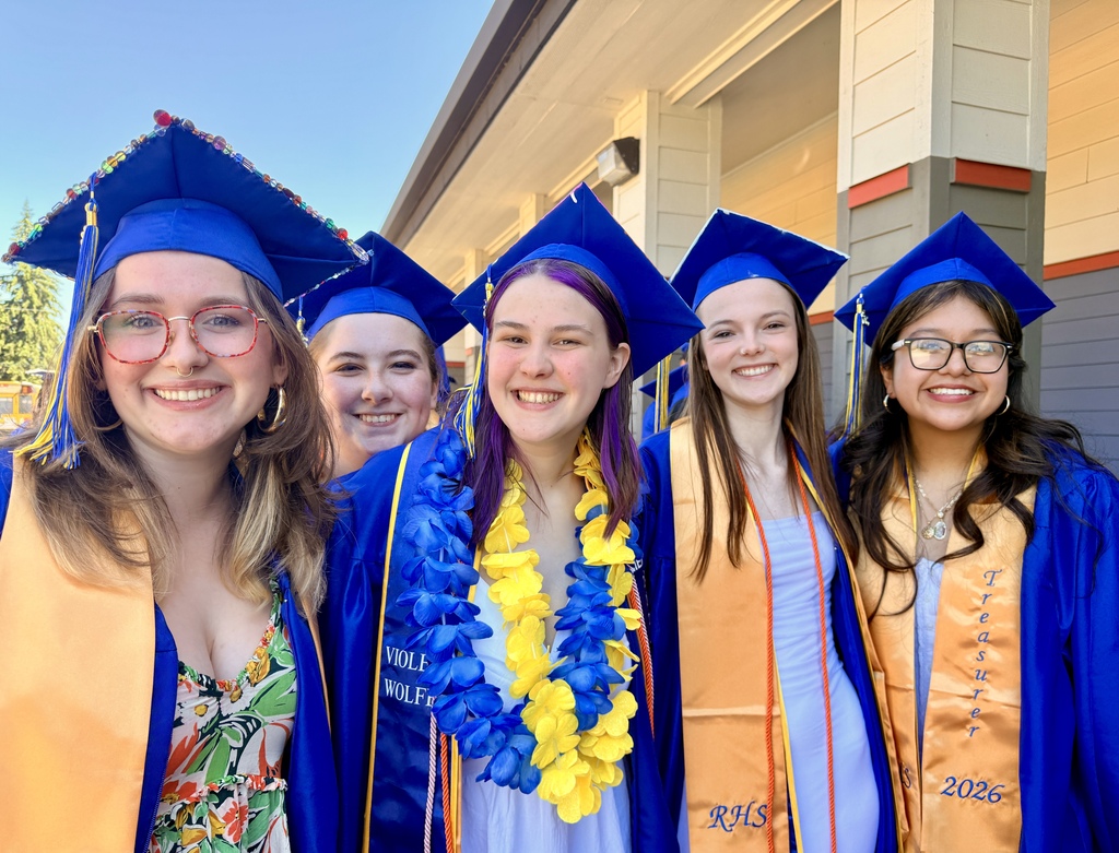 Five girls in blue caps and gowns smile at the camera outside a building. They wear necklaces and leis.