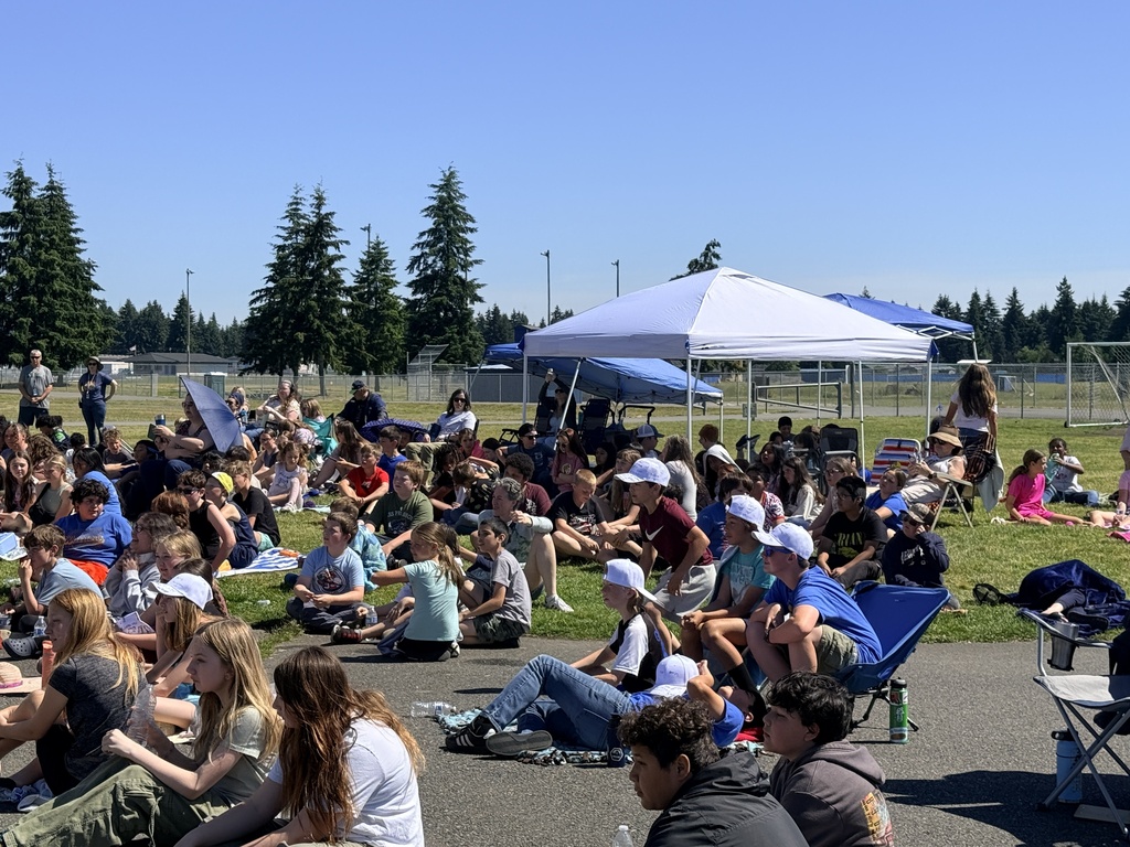 Many people seated on grass, some under umbrellas, and trees in the background.