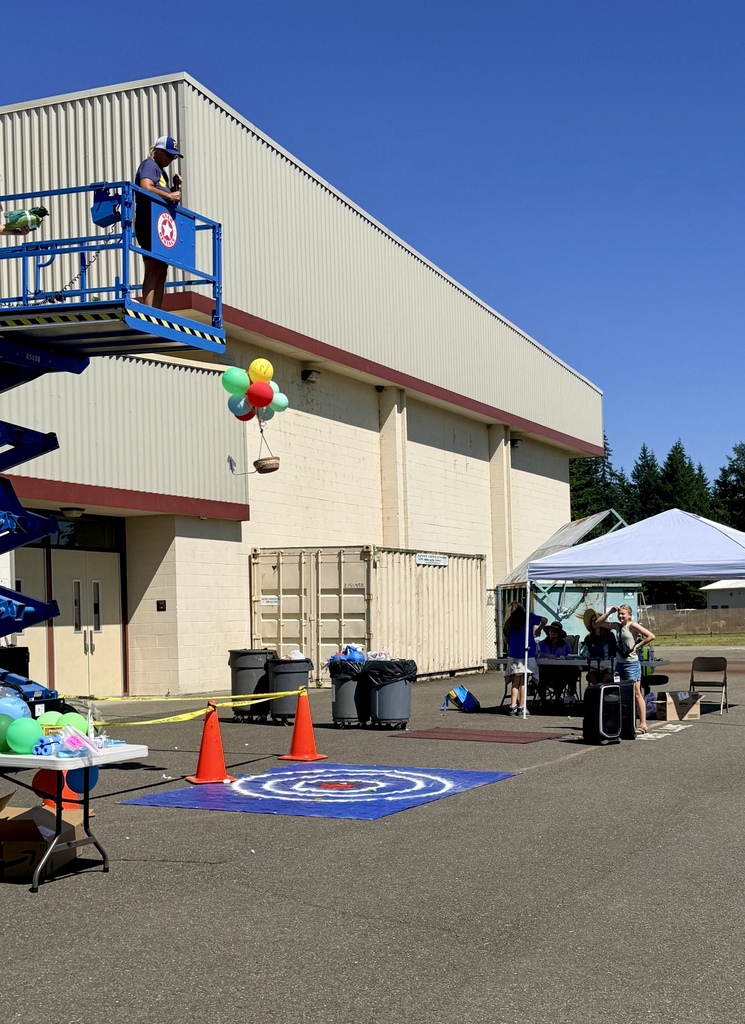 An event with balloons in air, a person on a platform, and people under a tent. Orange traffic cones and a yellow caution tape.