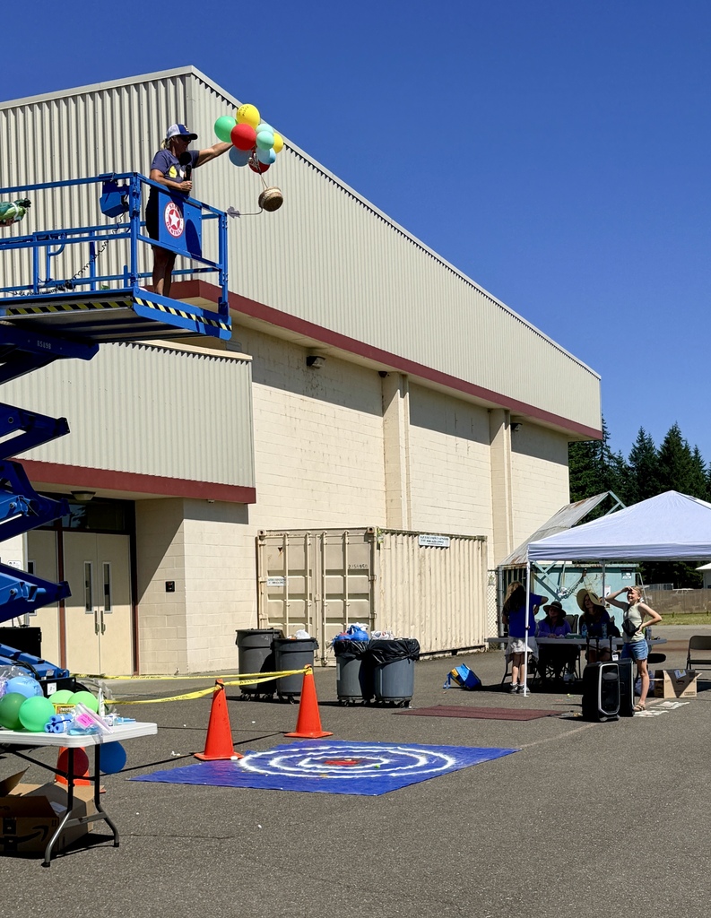 Person on platform dropping balloons; white tent; orange traffic cones; tables with items; building with doors.