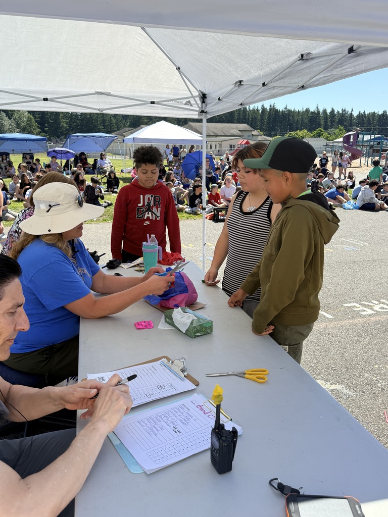 People under a tent, including adults in hats, with one holding a clipboard and writing, and another holding a pen.
