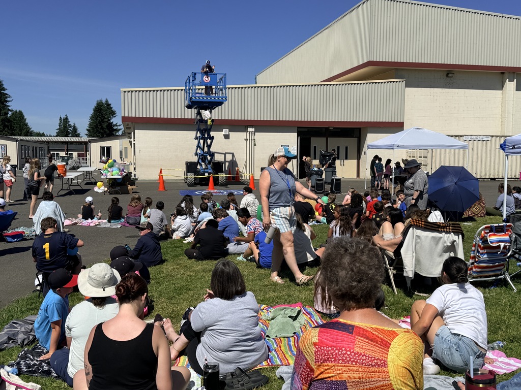A crowd sits on the grass in front of a building with people standing and an elevated platform.