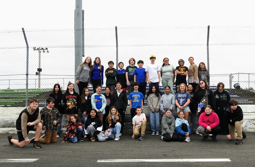 Group of individuals in various attires posing for a photo in front of a fence at the race track.