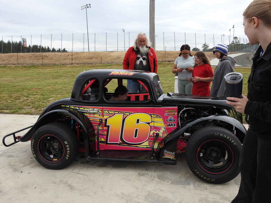 Several people stand around a vintage car with the number 16 on it. An open field and fence are in the background.