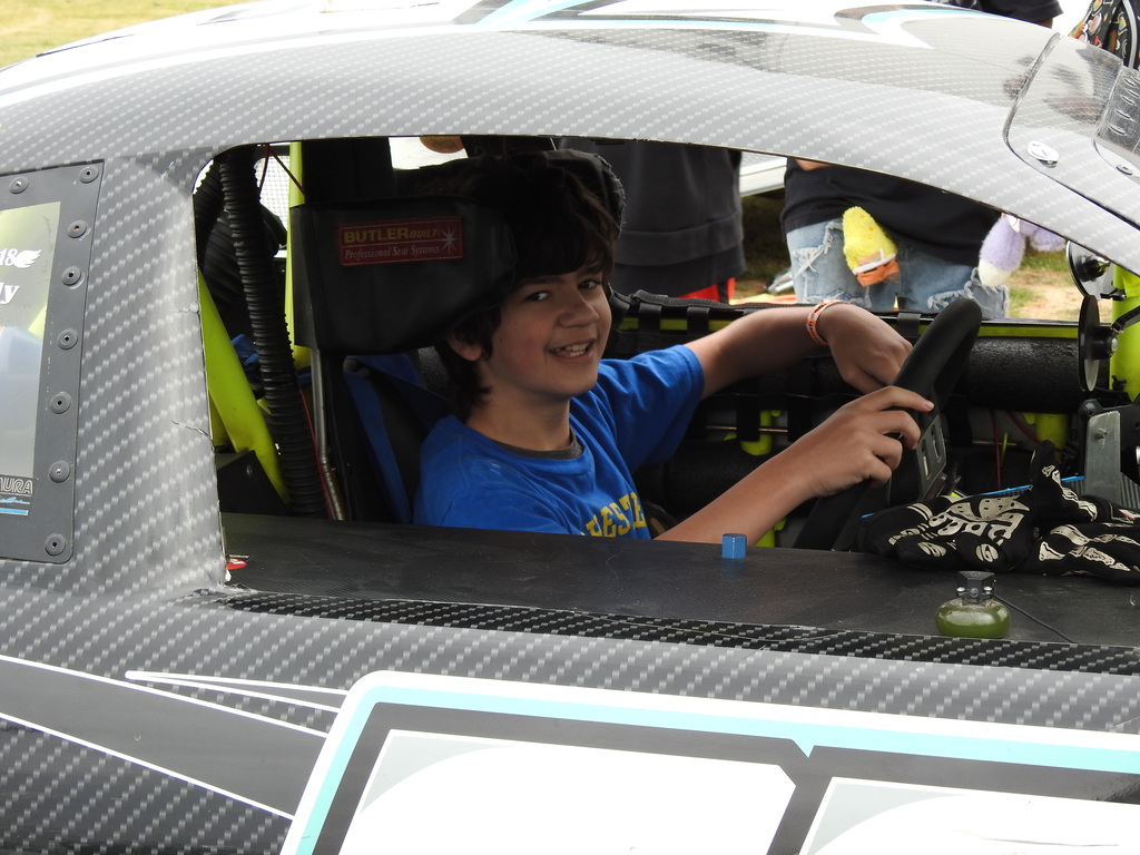 A boy smiles inside a racing car, holding the steering wheel. He sits in the driver's seat with a black and green seat.