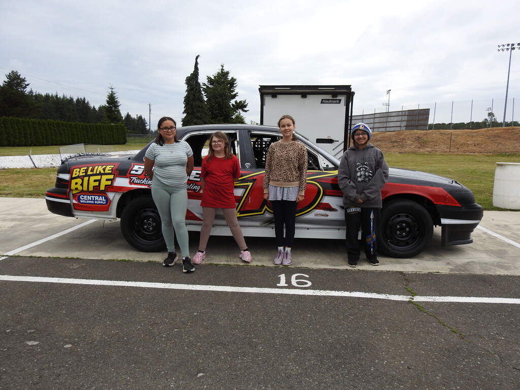 Four people stand in front of a car with text and numbers on the side. Behind them is a grassy area and a building.