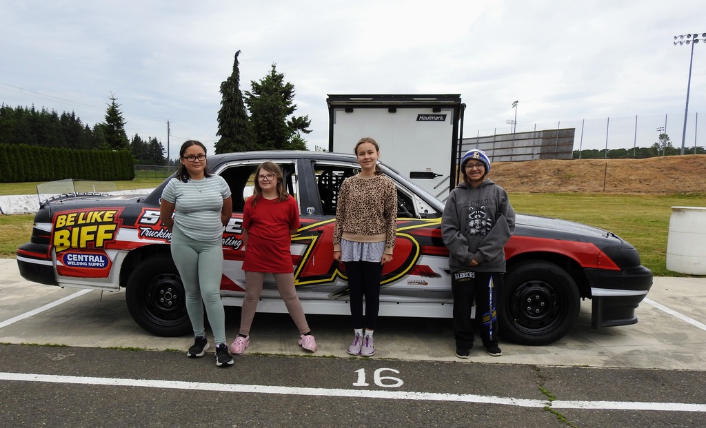 Four people stand in front of a black and red race car with white letters and numbers.