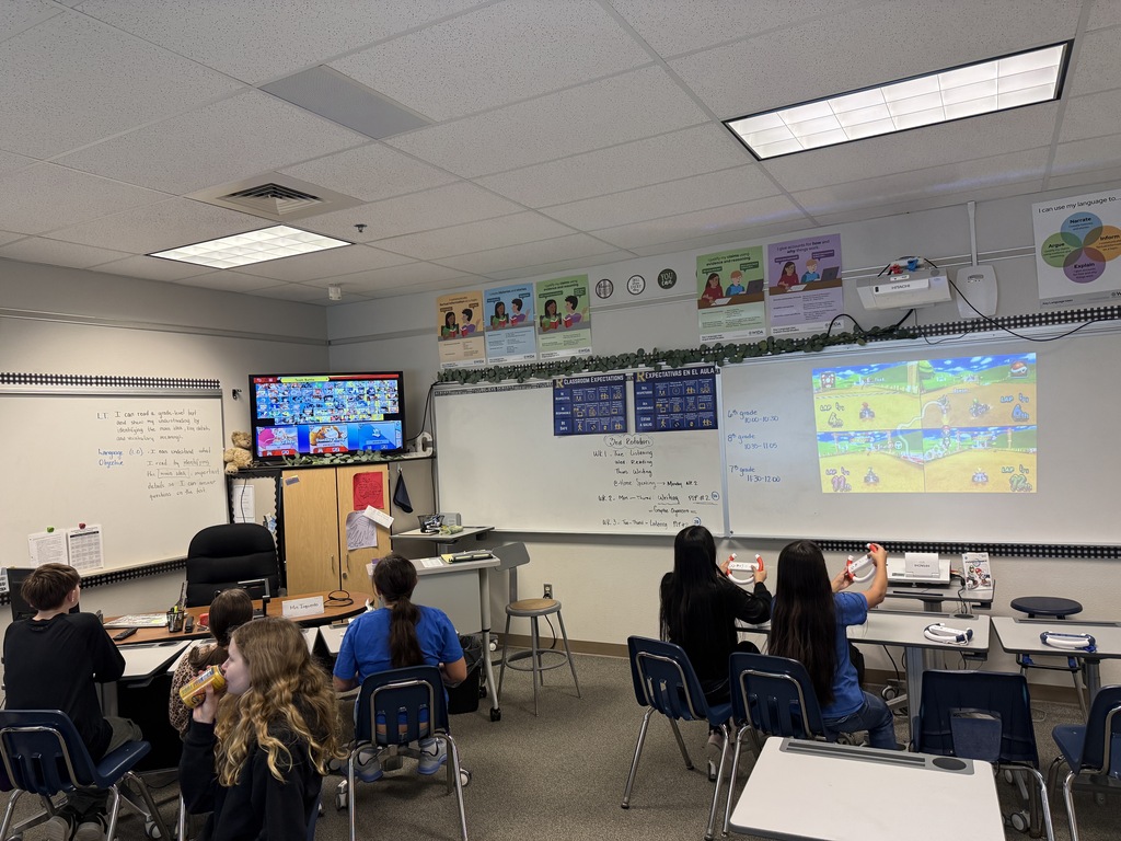 A classroom with chairs, tables, and students seated. Whiteboards and monitors on the wall, and a projector screen.