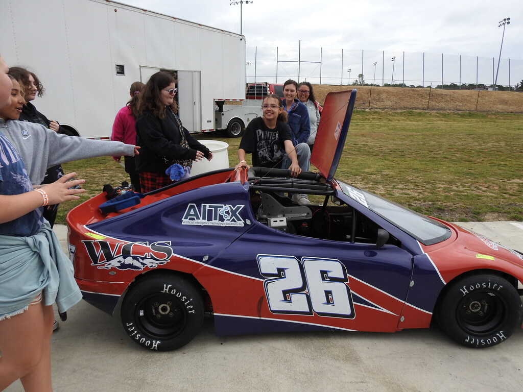 People gather around a race car. The car is red, blue, and white with the number 26 on the side.