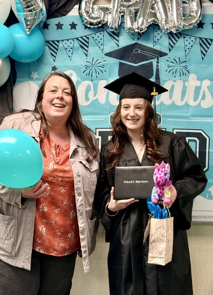 Two women pose in front of a graduation backdrop with balloons. One holds a blue balloon, the other a diploma and a gift bag.