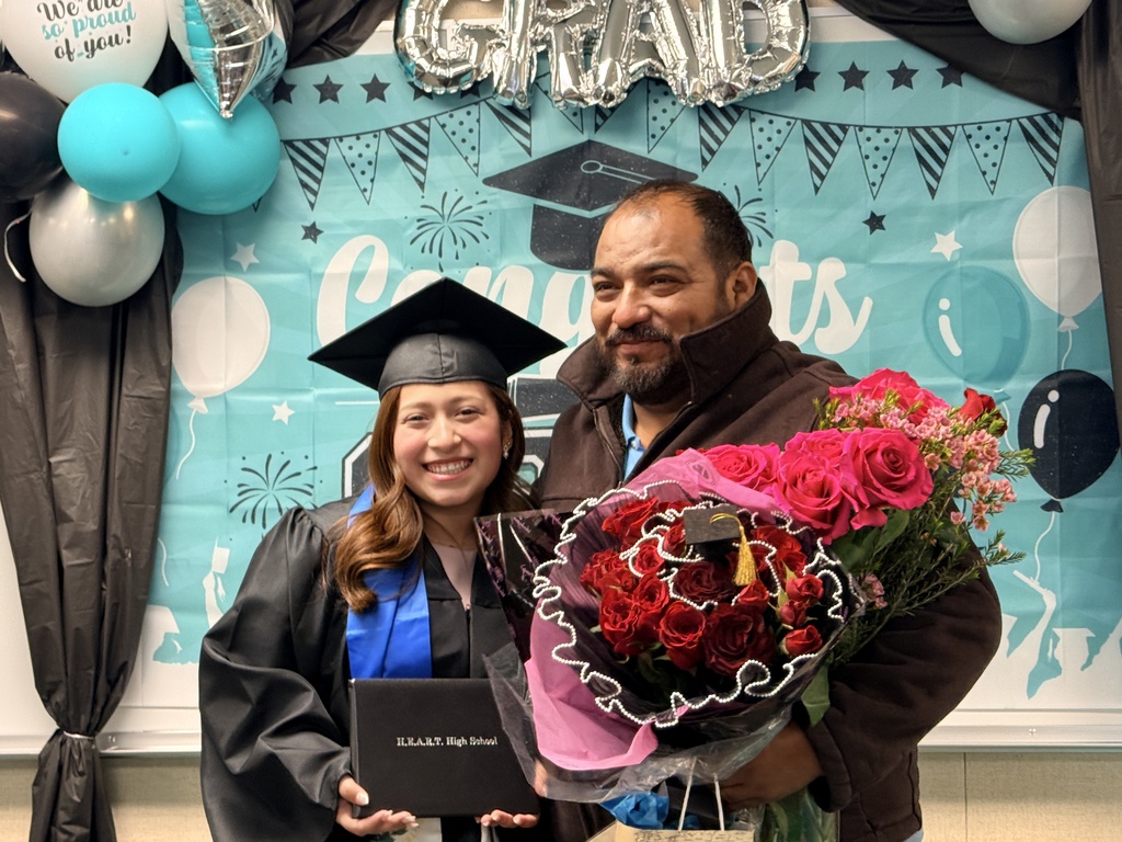 A woman in a graduation gown holds a diploma. A man holds flowers. Behind them is a banner with balloons and a graduation cap.