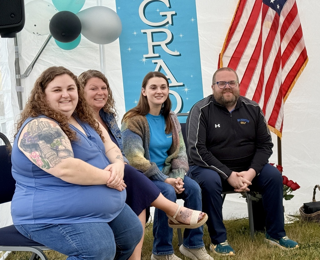 Four people sit in chairs under a tent. A blue banner reads "GRAD" and an American flag is nearby.