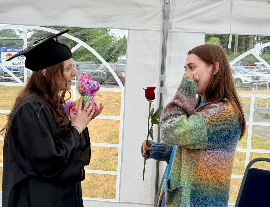 A woman in a graduation gown holds flowers while speaking to another woman in a colorful sweater. They are under a white tent.