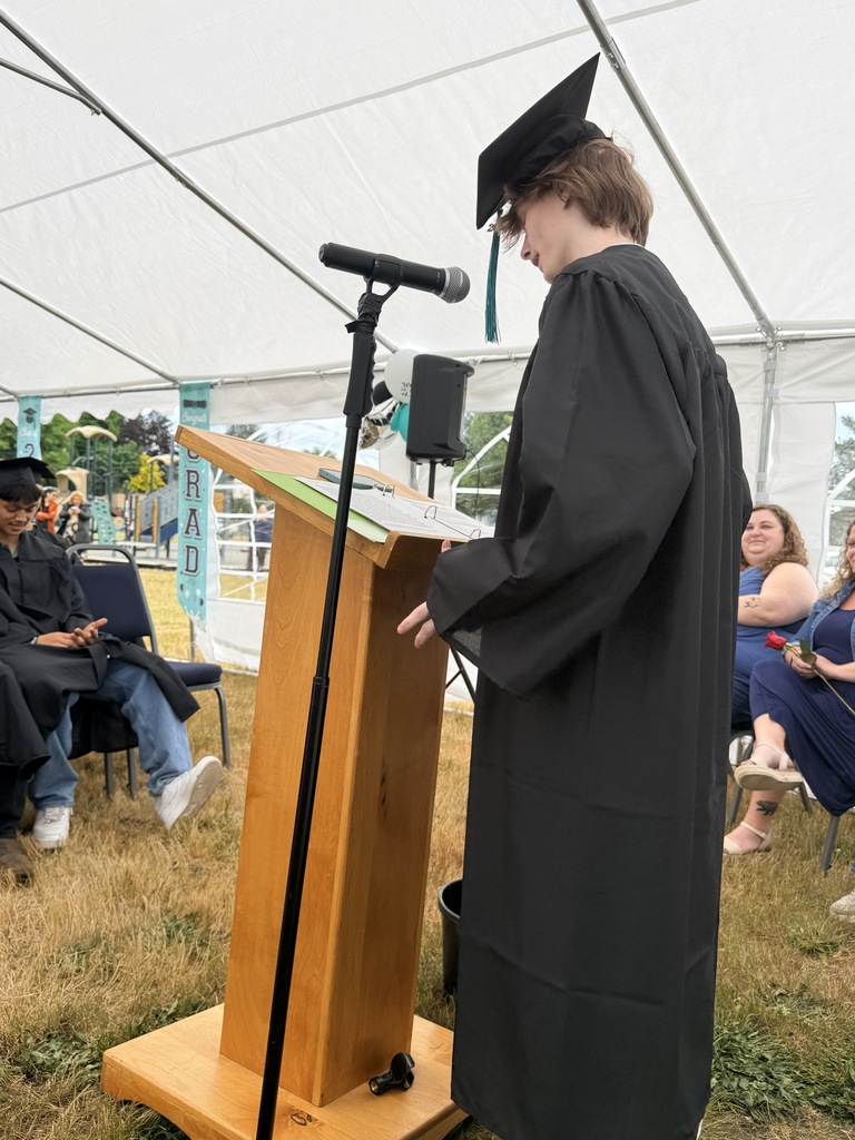 Person wearing a graduation gown standing at a podium with a microphone, reading from a book.