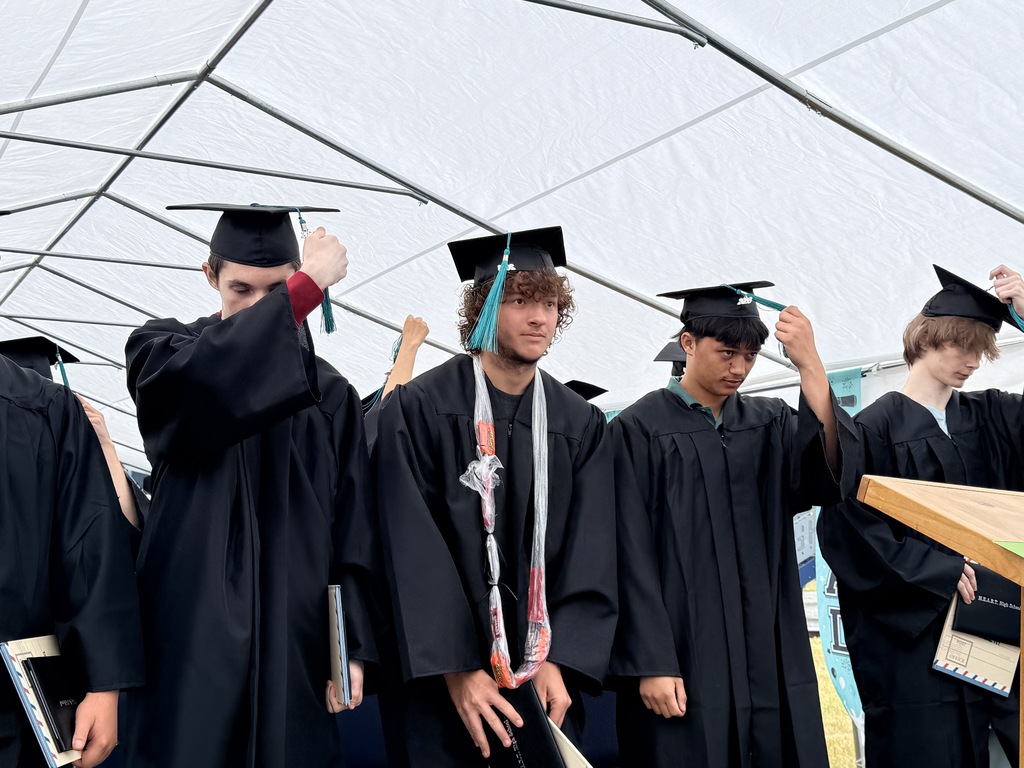 A group of people in black graduation gowns and hats under a white tent, some adjusting their hats.