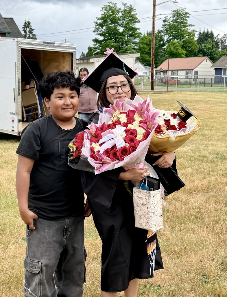 A person in a cap and gown holds flowers next to a boy in casual clothes. They stand in a grassy area with a white truck and houses in the background.