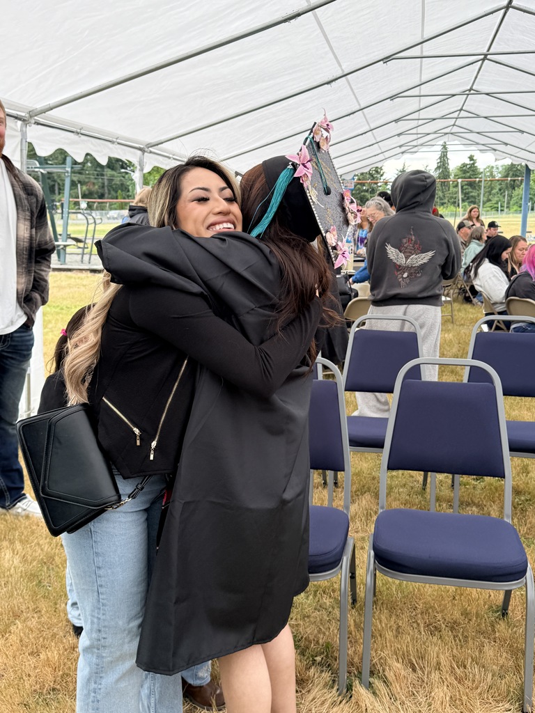 Two women hug under a tent, one wearing a black graduation cap. People sit in blue chairs in the background.
