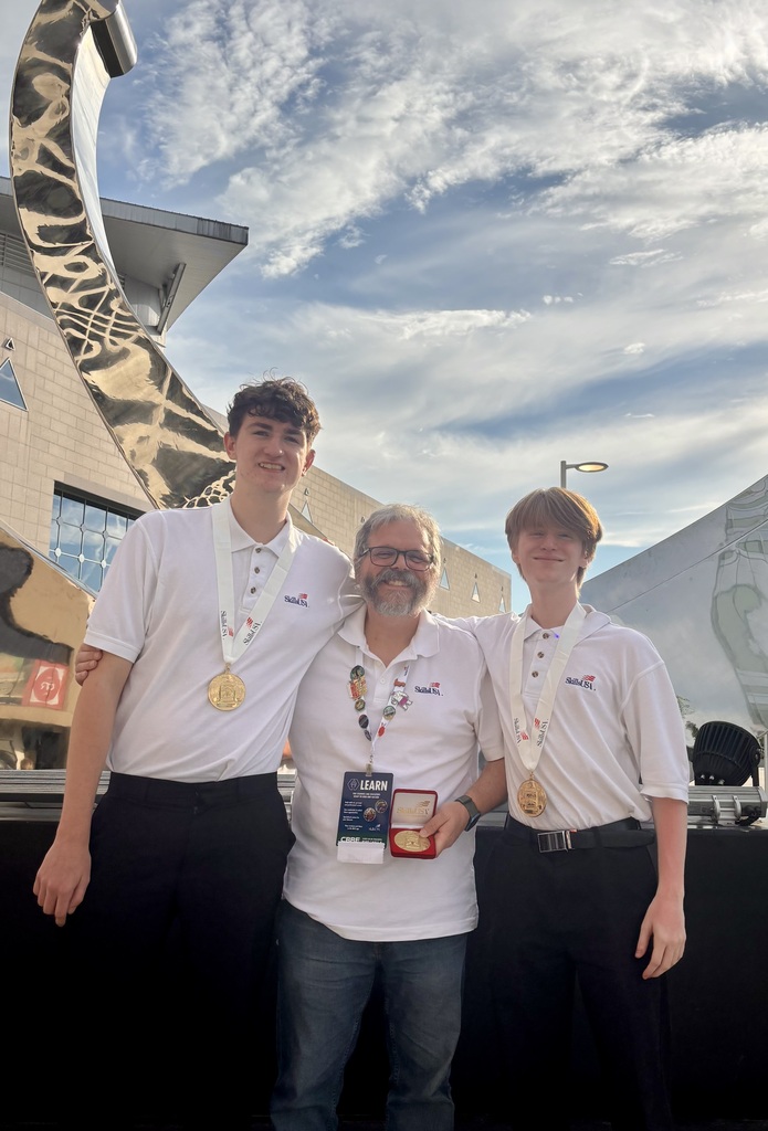 Three men, each wearing white shirts and black pants, stand together. The middle person has glasses and a watch. They wear medals and stand before a building.