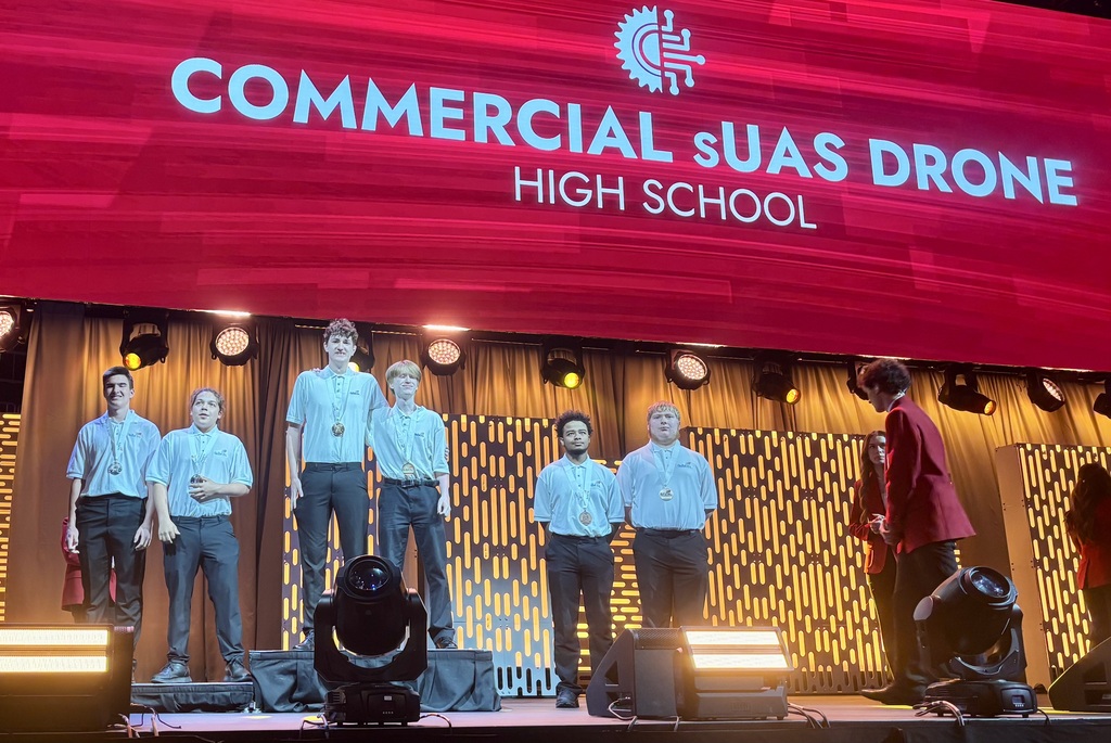 Group of people on a stage with a banner behind them that reads "Commercial SuAs Drone High School".