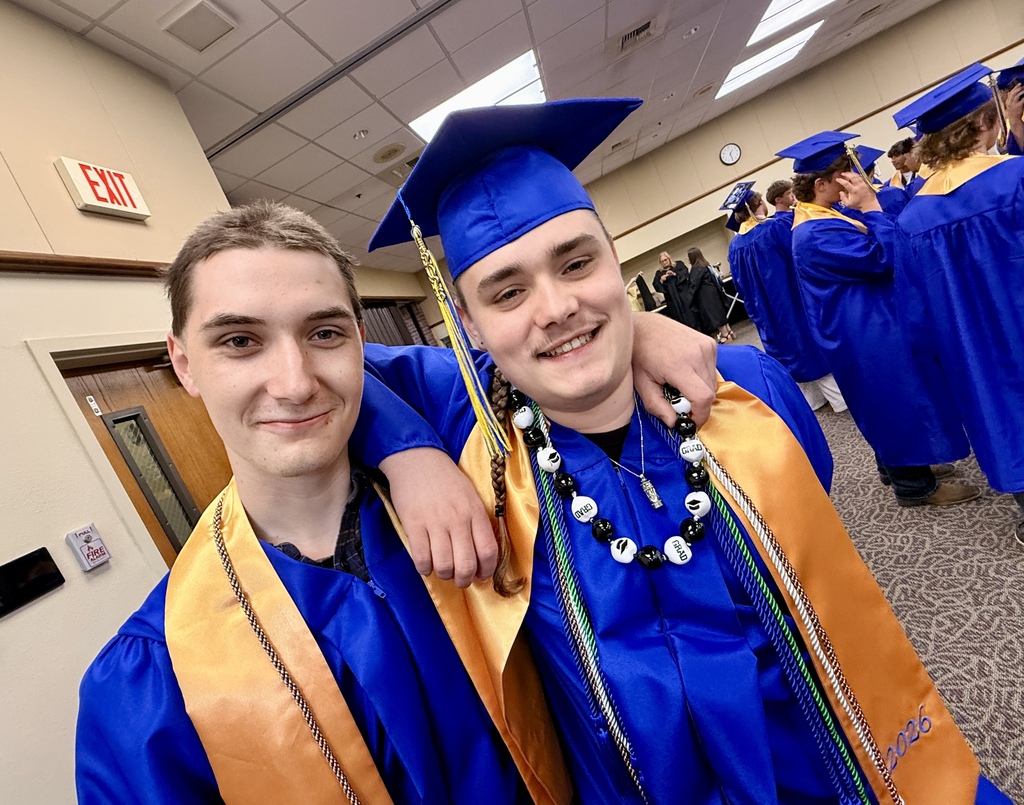 Two men in blue gowns and gold stoles smile at the camera. One wears a blue graduation cap and a necklace.