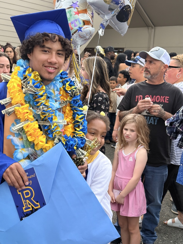 A young man in a cap and gown, wearing a lei, holds a gift bag and stands with two children. A crowd of people stands behind them, some wearing hats and glasses.