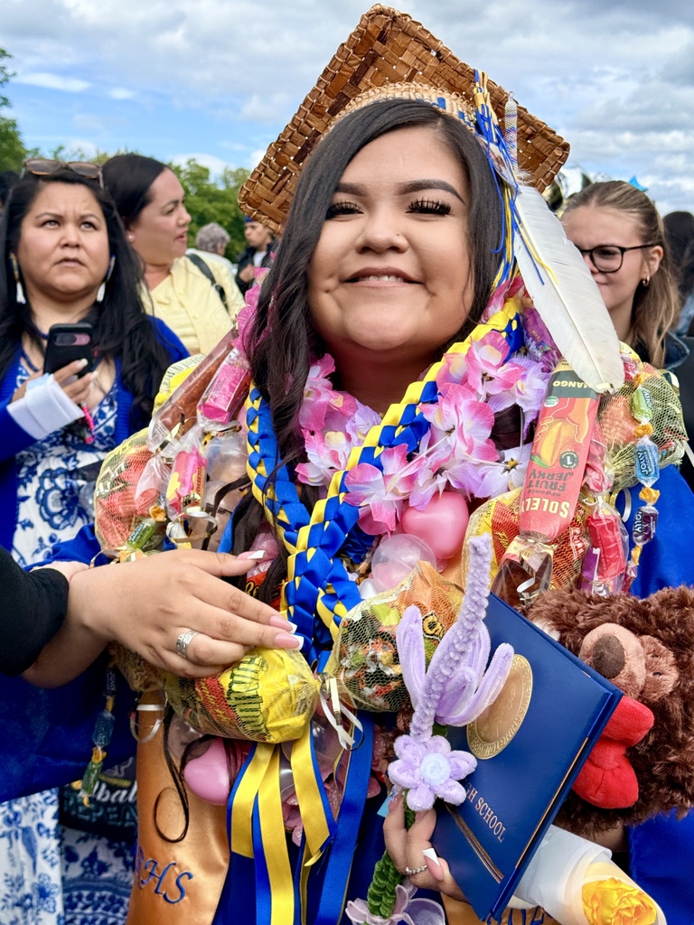 Woman in blue graduation gown, smiling. Wearing lei, holding cap, book, and toys. People and trees in the background.