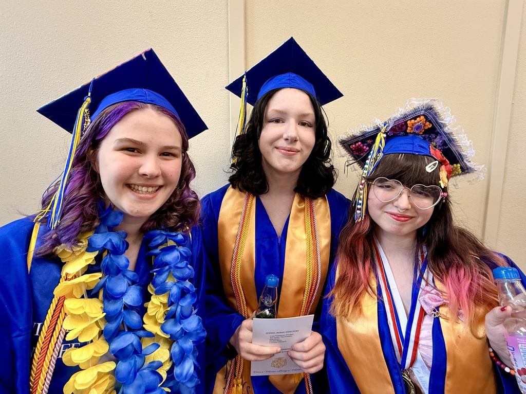 Three women in graduation robes with blue tassels and blue caps. Two hold certificates. The woman on left has a lei.