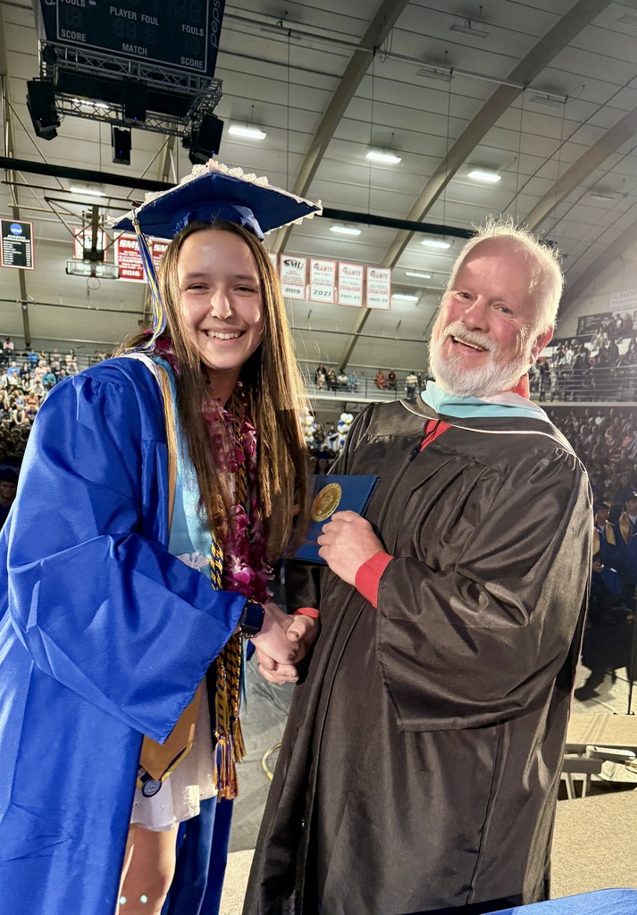 A woman in a blue gown shakes hands with a man in a black gown, both smiling. They are at a graduation ceremony.