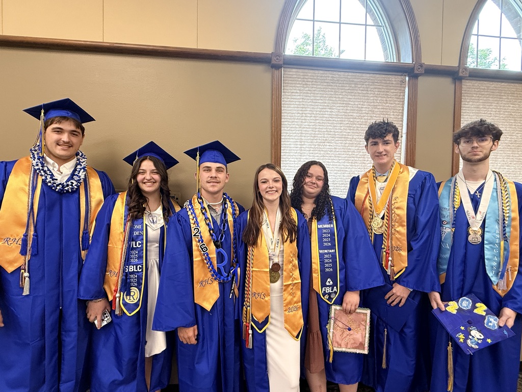 A group of eight students in blue graduation gowns and caps pose in front of arched windows.