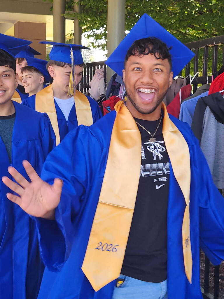 Graduates in blue caps and gowns, one smiling and waving. Background shows greenery and a fence.