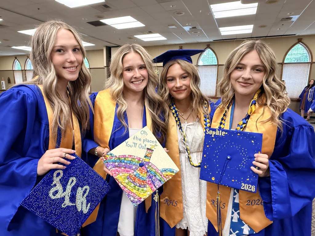 Four women in graduation gowns pose with decorated caps. The middle two wear blue and gold sashes.