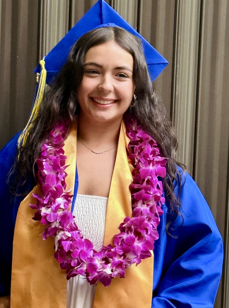 Woman in blue graduation cap and gown, smiling. Wearing lei of purple and yellow flowers around neck.