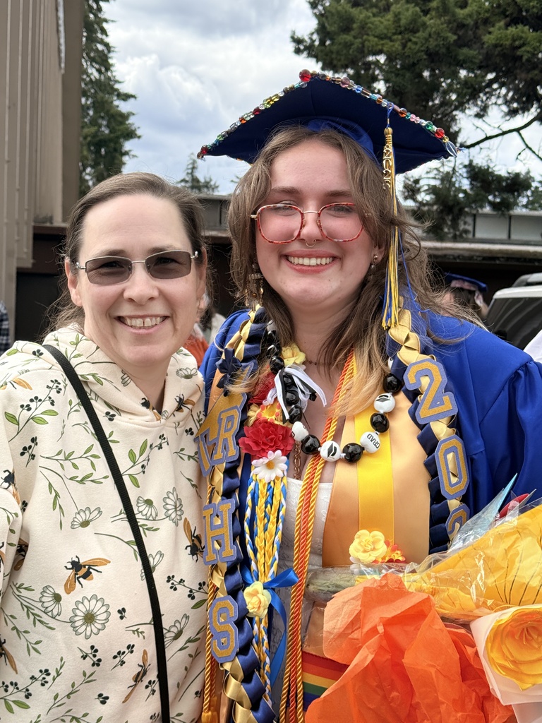 Two women smiling, one in a floral top and glasses, the other in a blue cap, gown, and necklace.