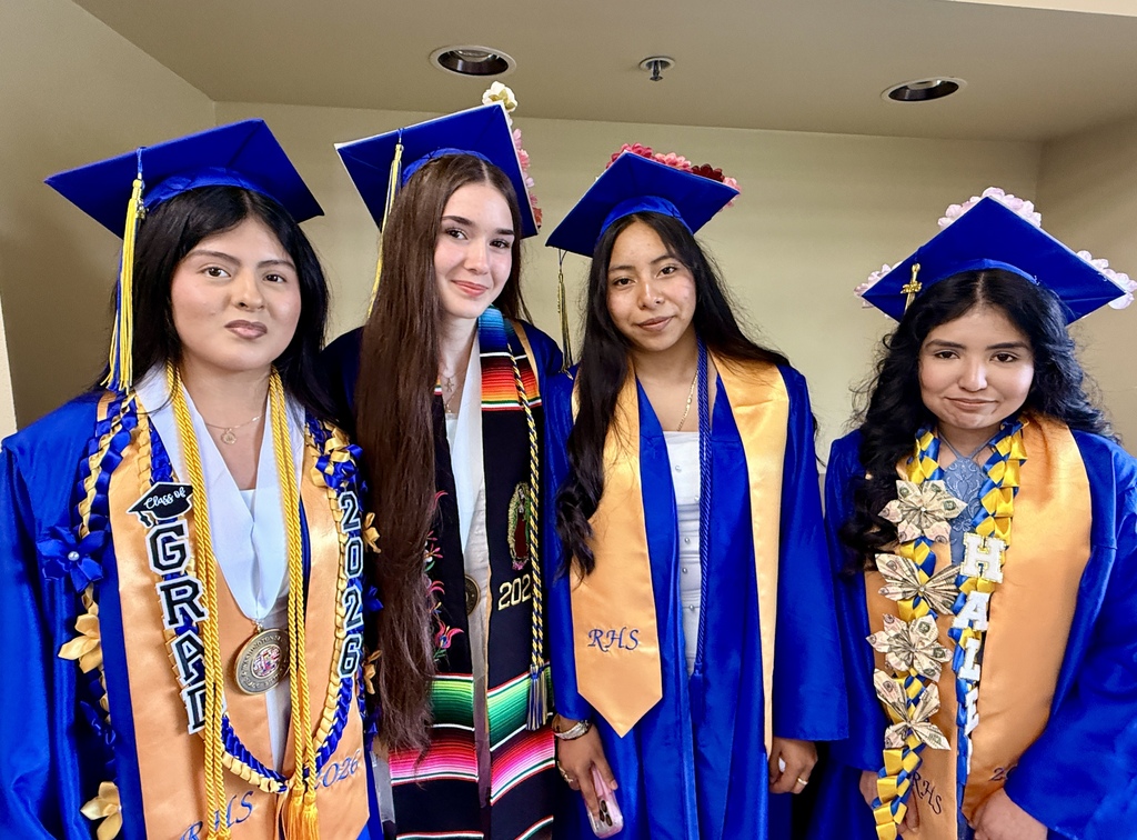 Five women in graduation gowns and hats pose for a photo in a hallway. Two wear blue, and three wear blue and gold.