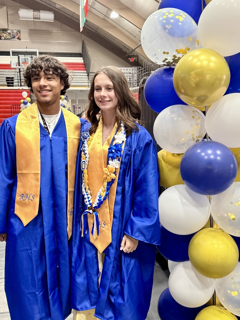 Two people in blue and gold graduation robes stand near a tall wall of blue and white balloons.