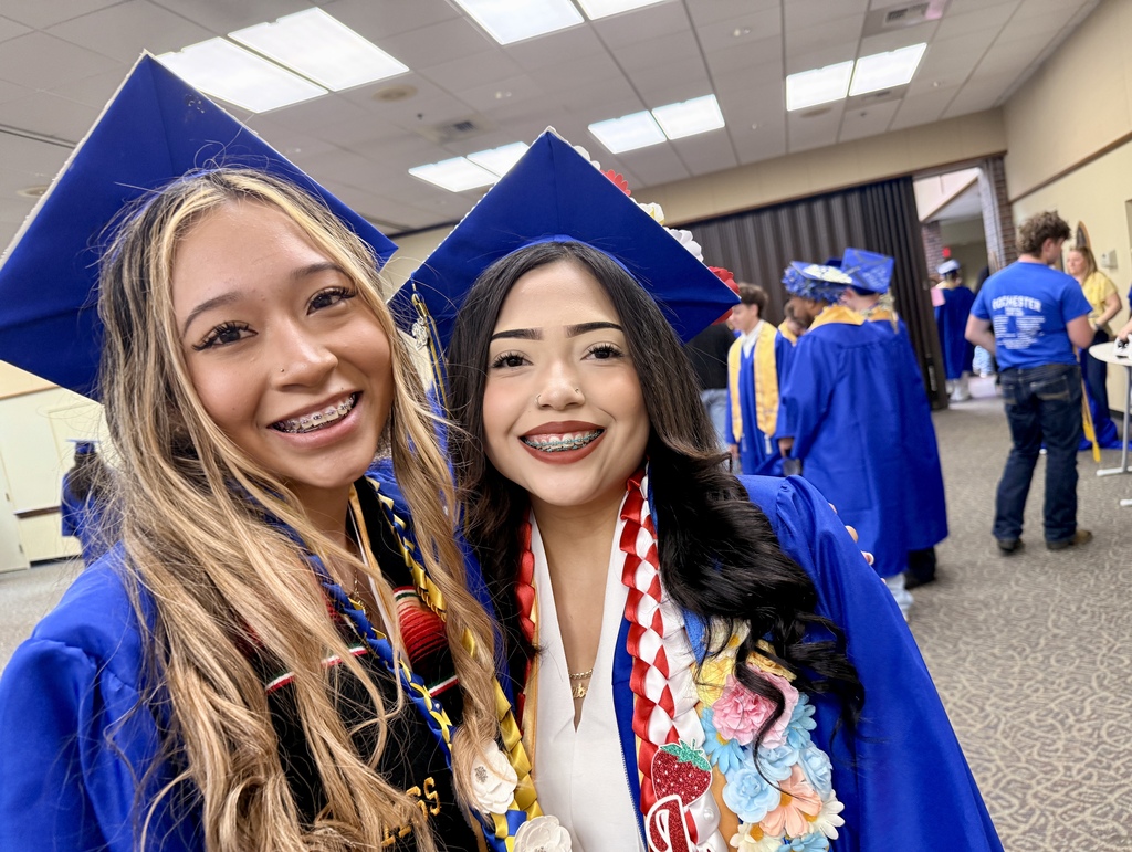 Two women in blue graduation gowns smile at camera. One has a red and white sash.