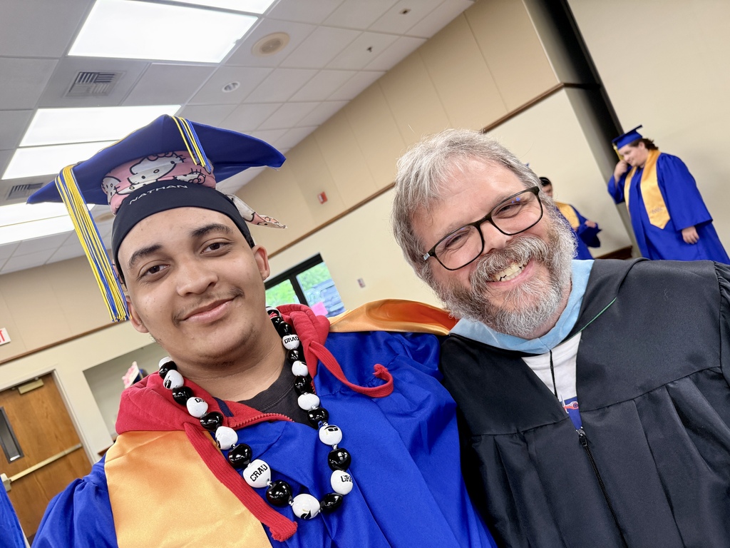 A man in a graduation gown stands next to a man in a graduation cap and a necklace.