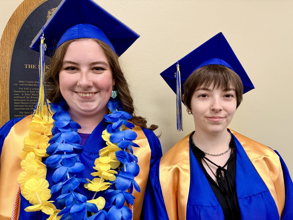 Two women in graduation gowns, one wearing a blue and yellow lei, stand next to each other.