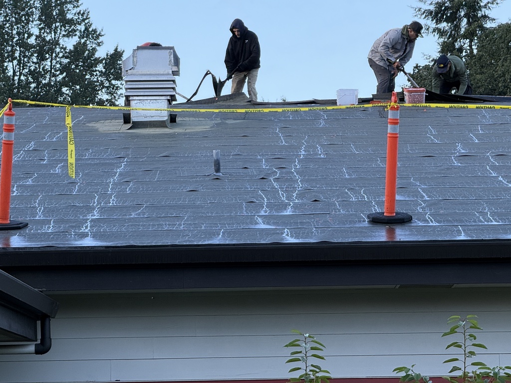Three workers on a roof with caution tape. One uses a sprayer, another holds a tool. Orange cones are present.