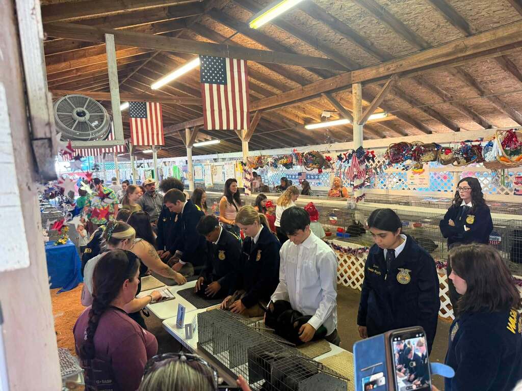 A group of people in a covered area with wooden ceiling beams. Some wear uniforms and hats, others carry bags.
