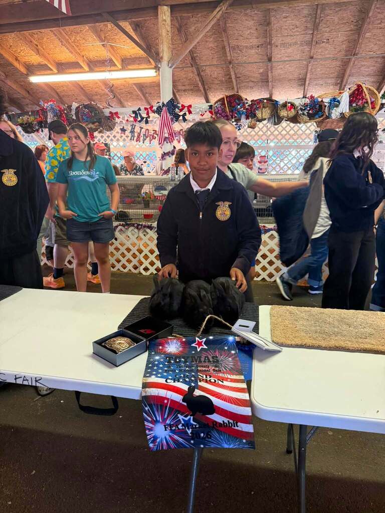 A child stands behind a table with an American flag banner and other items. People move in a room with a white fence.