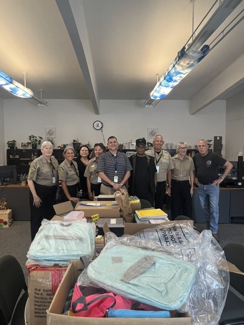 Several people stand behind a table with many boxes, some containing backpacks and clothes, in a room.