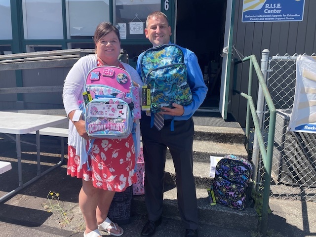 A man and a woman stand in front of a building, both holding backpacks. The woman wears a floral dress, and the man a suit.