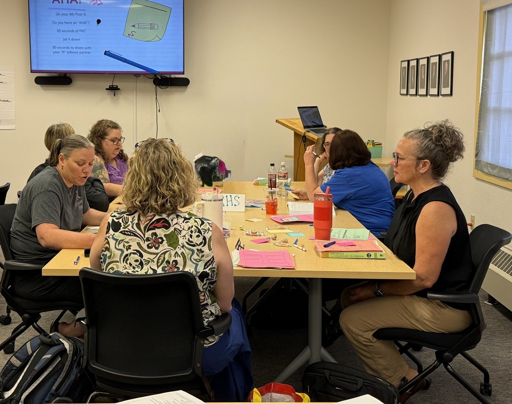 A group of people seated around a table in a classroom, facing a projector screen.